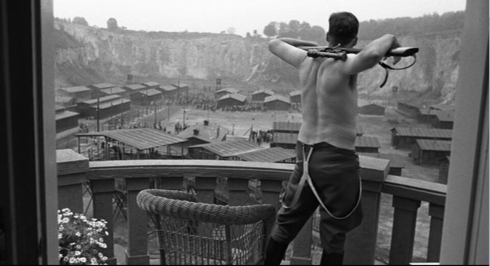 Zdjęcie przedstawia Gӧtha stojącego na balkonie, w samych wojskowych spodniach. Mężczyzna trzyma na karku karabin. Patrzy się na plac obozowy pełen baraków.