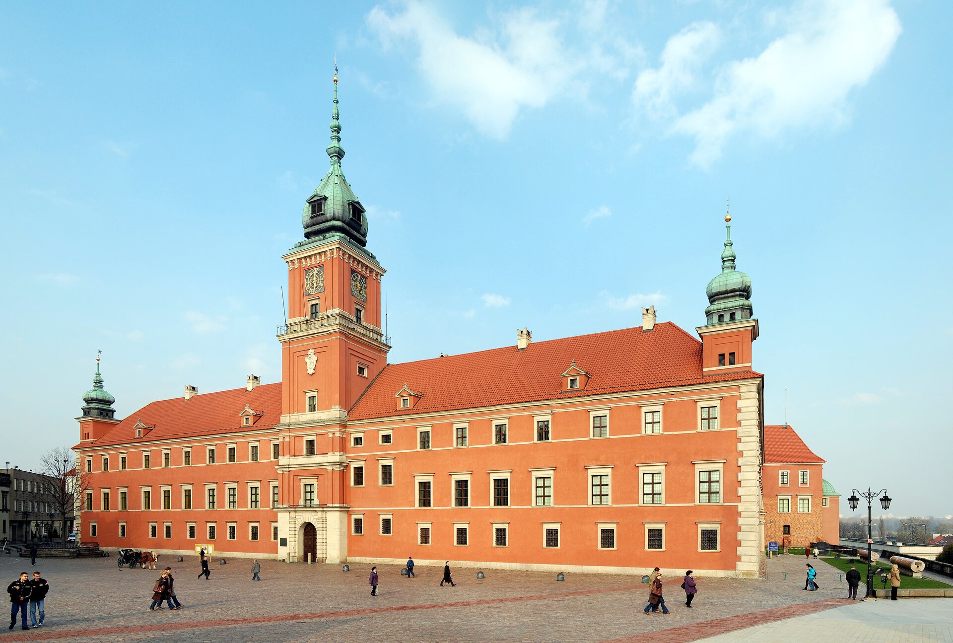 Zdjęcie przedstawia zamek Królewski w Warszawie. Budynek zbudowany jest z czerwonej cegły. Krawędzie szczytowych ścian budynku ozdobione są białymi cegłami. W centralnej części znajduje się wysoka wieża. Z jej czterech stron widoczny jest zegar. Na szczytach budynku znajdują się dwie, niższe wieże. Okna na pierwszym i drugim piętrze są wyższe, niż okna na parterze. Na placu przed zamkiem przechadzają się ludzie. 