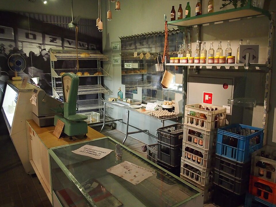 Zdjęcie przedstawia wnętrze sklepu spożywczego. Na ladzie stoi waga oraz kasa. Po lewej stronie za blatem stoi druciany regał z pojedynczymi bochenkami chleba. Na stoliku leży pusta tekturowa platonka po jajkach, w miseczce obok kilka jaj kurzych. Na wiszącej półce ustawione obok siebie kilkanaście jednakowych, szklanych butelek z octem oraz słoiczki z musztardą. Pod pułkami plastikowe skrzynki z pustymi butelkami po mleku oraz po oranżadzie. 