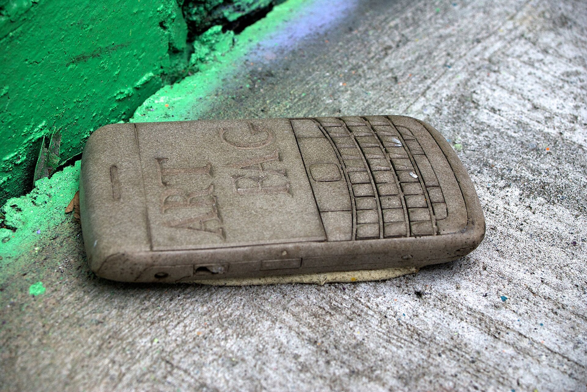 Ilustracja przedstawia rzeźbę Willa Colesa „Art Fag". Ukazuje wykonany z betonu telefon komórkowy. Nad klawiaturą znajduje się ekran, na którym został umieszczony napis: ART FAG. Telefon leży na chodniku przy betonowej ścianie pomalowanej na zielono.