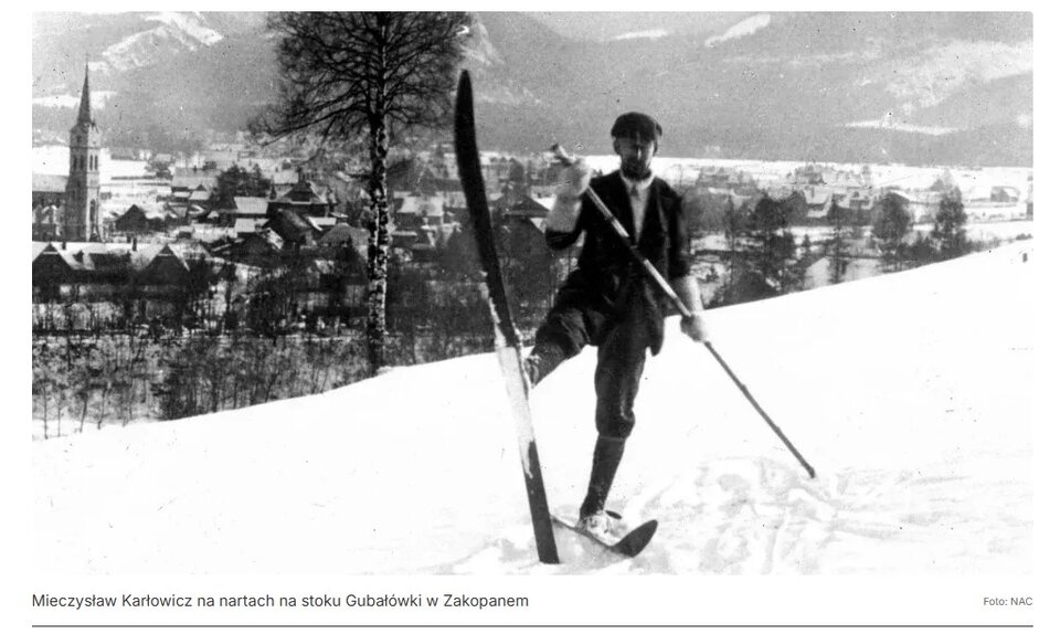 Stara, czarno‑biała fotografia przedstawia narciarza na stoku górskim. Narciarz stoi na jednej narcie, drugą nogę unosi, stawiając pionowo nartę do niej przypiętą. Podpiera się kijem narciarskim. W tle widoczna panorama miasta.