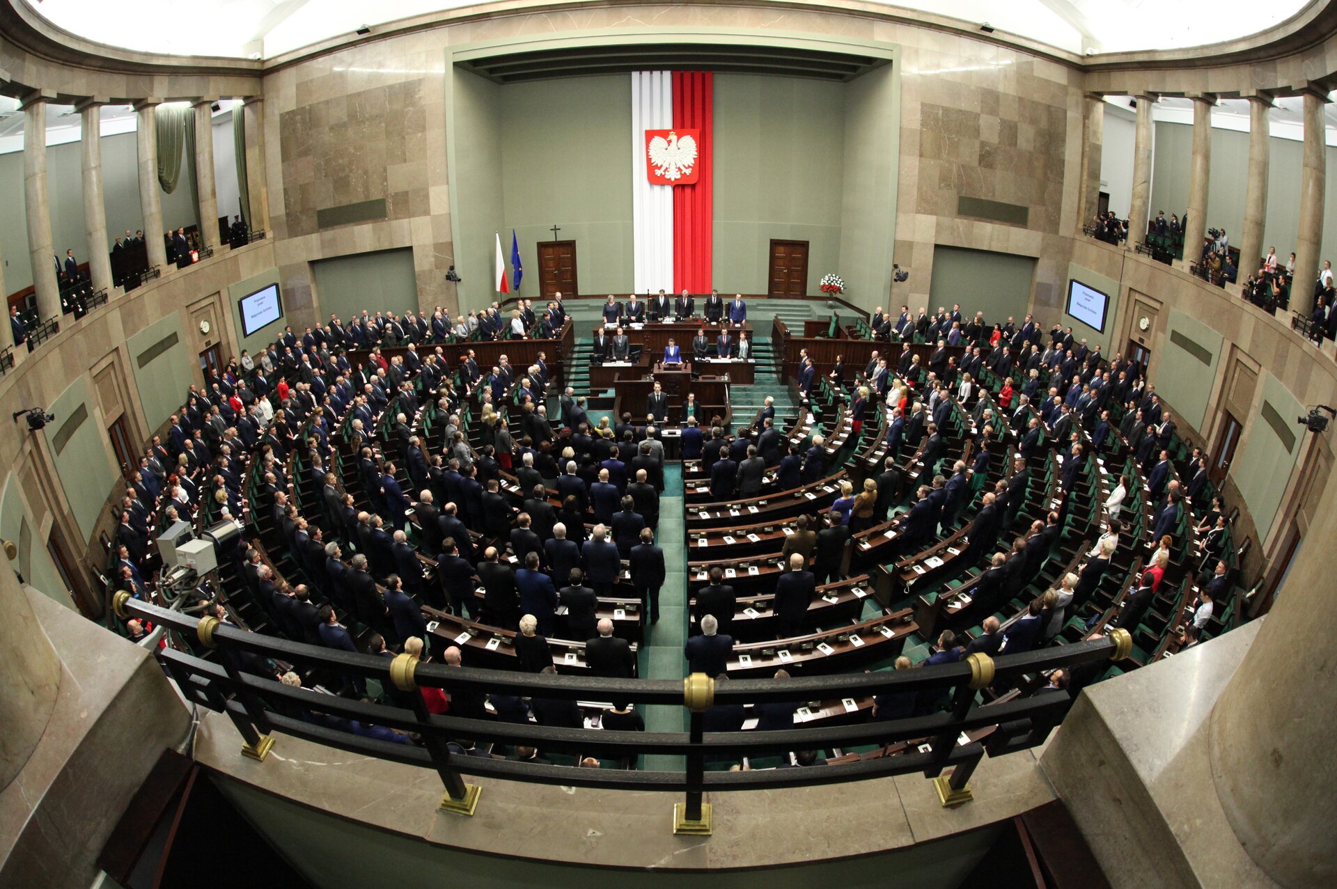 Zdjęcie przedstawia półokrągłą salę z ławami poselskimi. Zasiadają w niej liczni posłowie. Pośrodku sali znajduje się stół marszałka Sejmu i mównica. Za nimi, na ścianie wisi polska flaga.