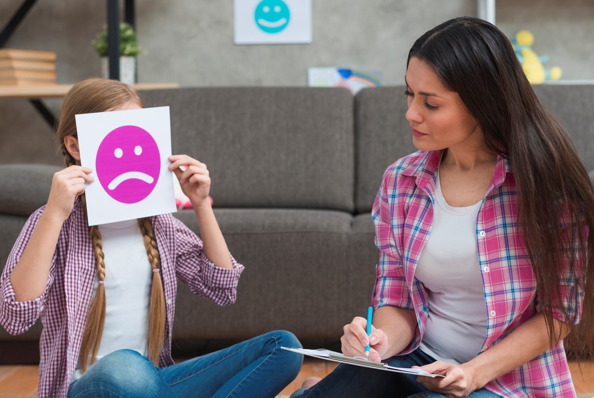 Zdjęcie przedstawia kobietę w młodym wieku siedzącą na kanapie z dziewczynką, która zakrywa twarz kartką, na której jest smutna buźka. Transkrypcja znajduje się poniżej.