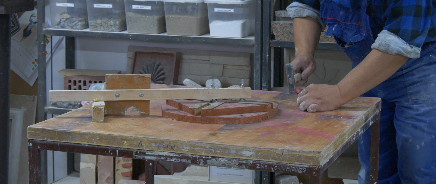 Zdjęcie przedstawia wykrój do profili łukowych. Na stole, przy którym pracuje pracownik w stroju roboczym, zostały zgromadzone drewniane wykroje w kształcie łuku różnej wielkości.