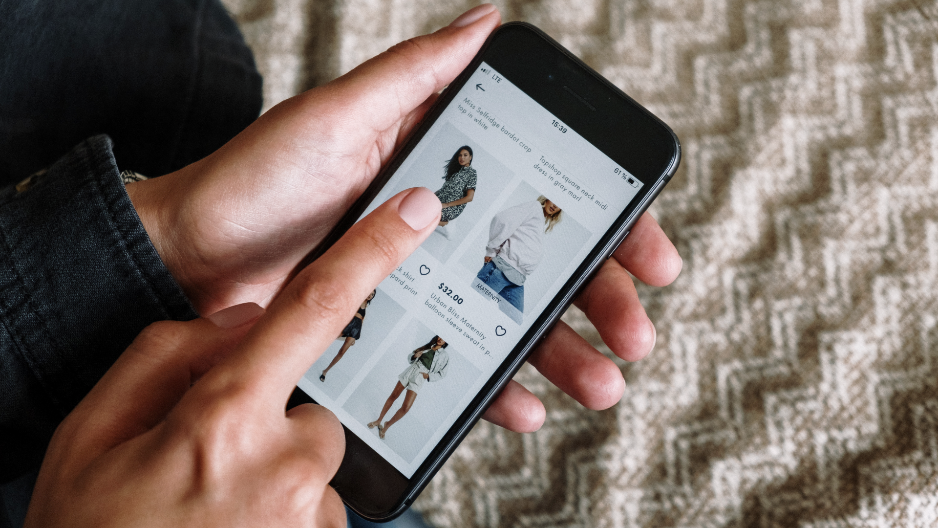 Zdjęcie przedstawia smartfon w rękach jakiejś osoby. Na ekranie wyświetlone są oferty sklepu z odzieżą.