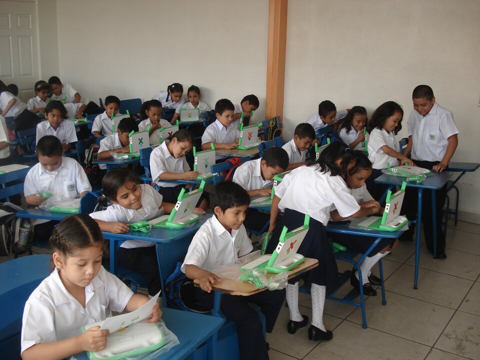 Zdjęcie przestawia klasę w szkole. Sala ma jasne ściany, z tyłu widać duże białe drzwi, na podłodze leżą duże prostokątne kafle. Przy pojedynczych niebieskich ławkach siedzą uczniowie, małe dzieci ubrane w jednakowe uniformy - dziewczynki mają ciemne obuwie, białe podkolanówki, ciemną spódnicę i białe koszule na krótki rękaw, chłopcy mają na sobie białe koszule i ciemne spodnie. Większość dzieci siedzi w swoich ławkach i pracuje przy użyciu biało‑zielonych laptopów. Pojedynczy uczniowie stoją przy ławkach swoich kolegów lub koleżanek, rozmawiając z nimi o swojej pracy.