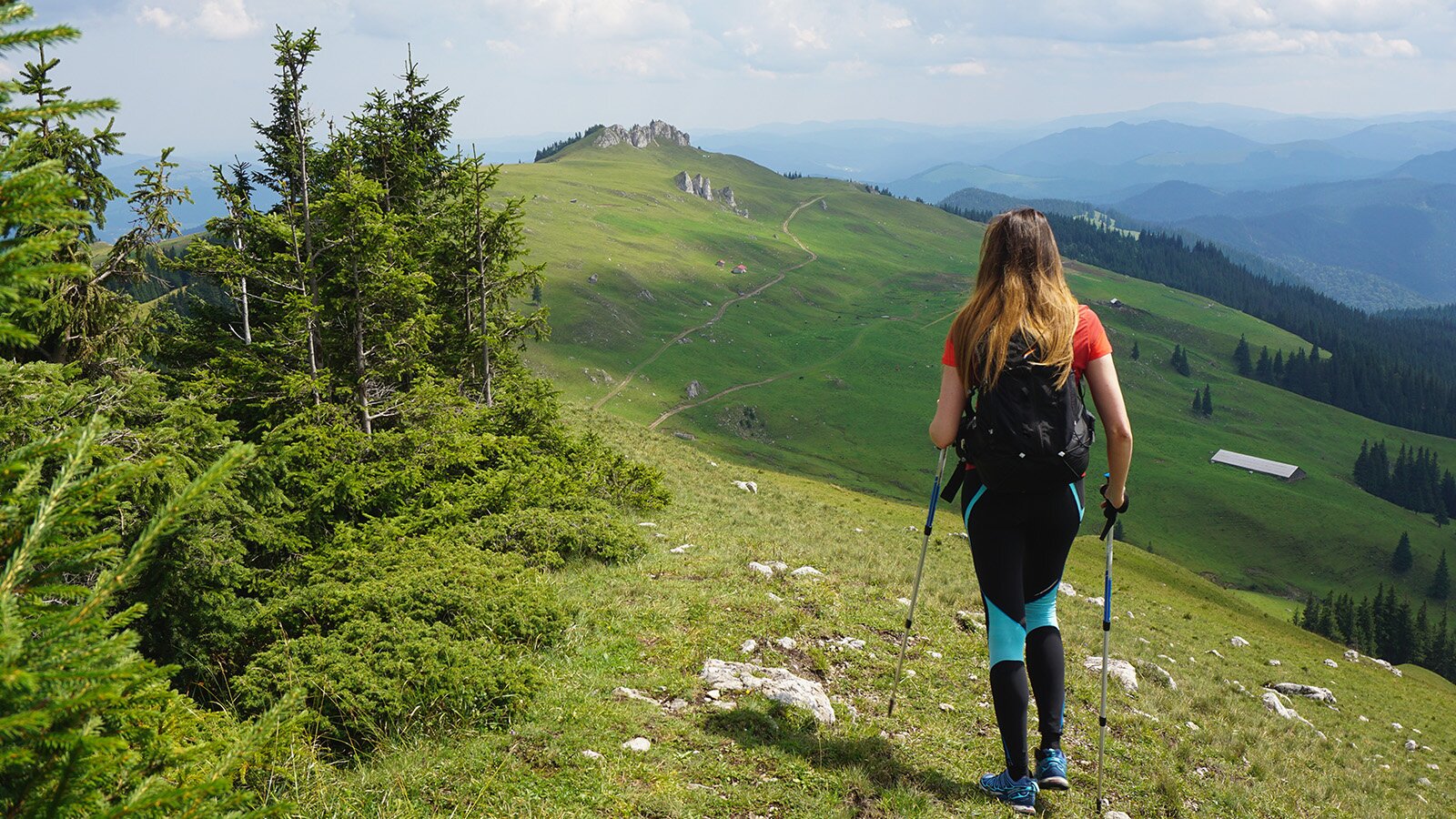 Zdjęcie przedstawia kobietę ubraną w strój sportowy, która trzyma w dłoniach kijki do marszu. Widoczne są pagórki oraz droga, po której idzie kobieta. 