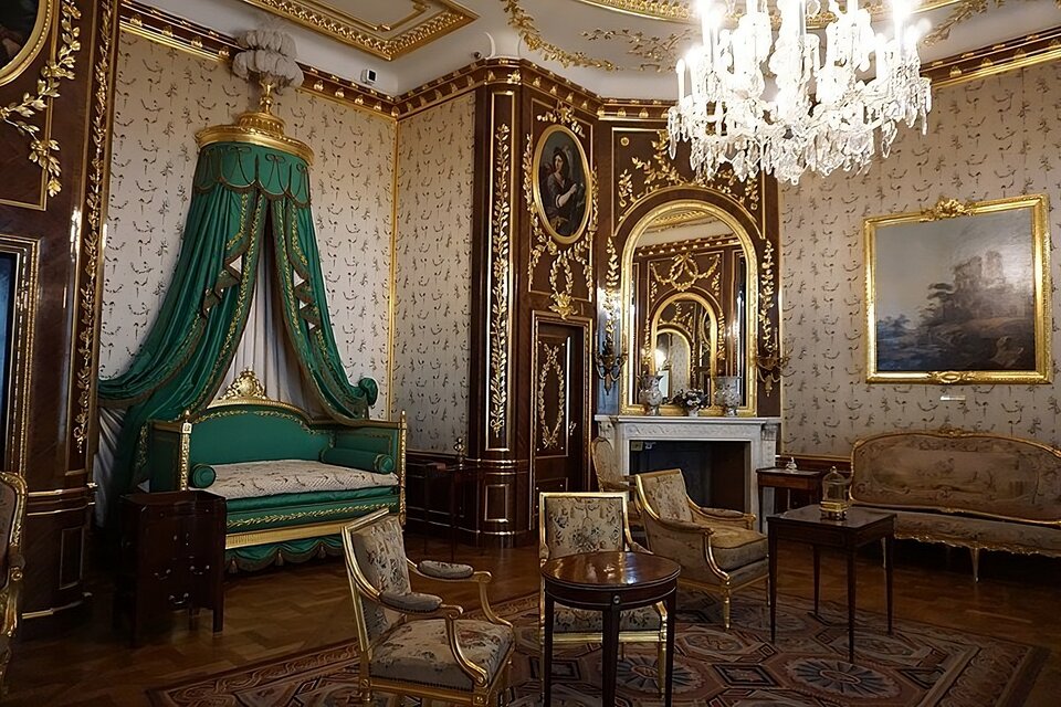 Zdjęcie przedstawia sypialnię Stanisława Augusta Poniatowskiego. Jej ściany pokrywają jasne tkaniny przedzielone co kilka metrów ciemną boazeria ze złoconą dekoracją. Po lewej stronie umieszczone jest łoże tureckie z baldachimem. Po jego bokach drzwi. Obok drzwi jest biały, marmurowy kominek z umieszczonym nad nim lustrem. Po środku pokoju stoją meble: krzesła, sofy i stoliki kawowe. Na kominku stoją wazy, na ścianie po prawej wisi obraz. 