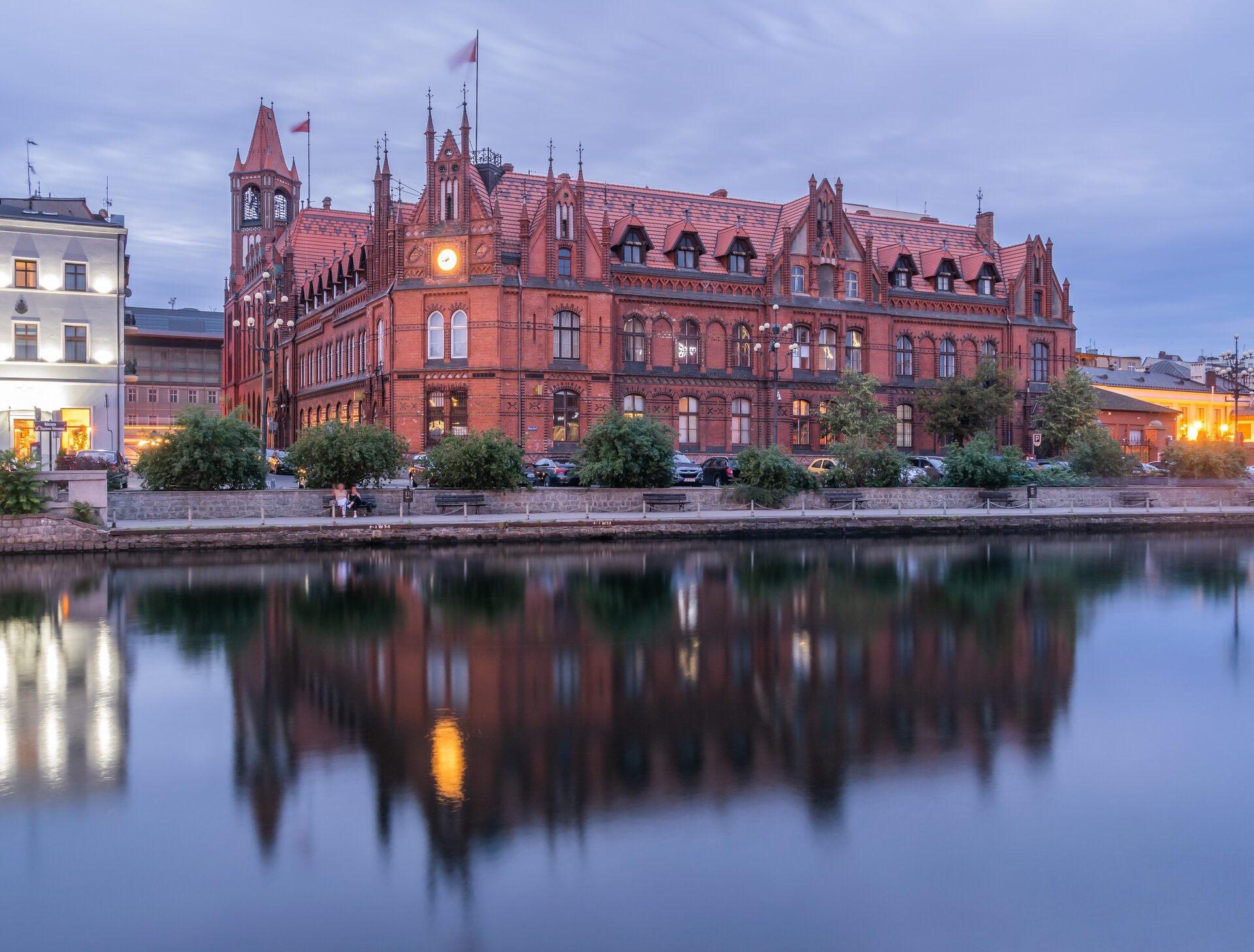 Fotografia przedstawia budynek Poczty Polskiej, który zbudowany jest z czerwonej cegły w stylu neogotyckim. Na lewej ścianie budynku znajduje się wieża - najwyższa jego część. W narożniku budynku znajduje się szczyt z zegarem. Wysokie, półokrągłe okna ustawione są w dwóch rzędach jedno nad drugim, a wyżej znajdują się jeszcze okna dachowe. Na dachu budynku umieszczono dwie flagi. Przy budynku biegnie alejka z ławkami, a na jednej z ławek siedzą dwie postacie. Alejka znajduje się przy brzegu rzeki. Obok rosną krzewy oraz drzewa. Dalej ukazana jest ulica oraz parking, na którym znajdują się auta.