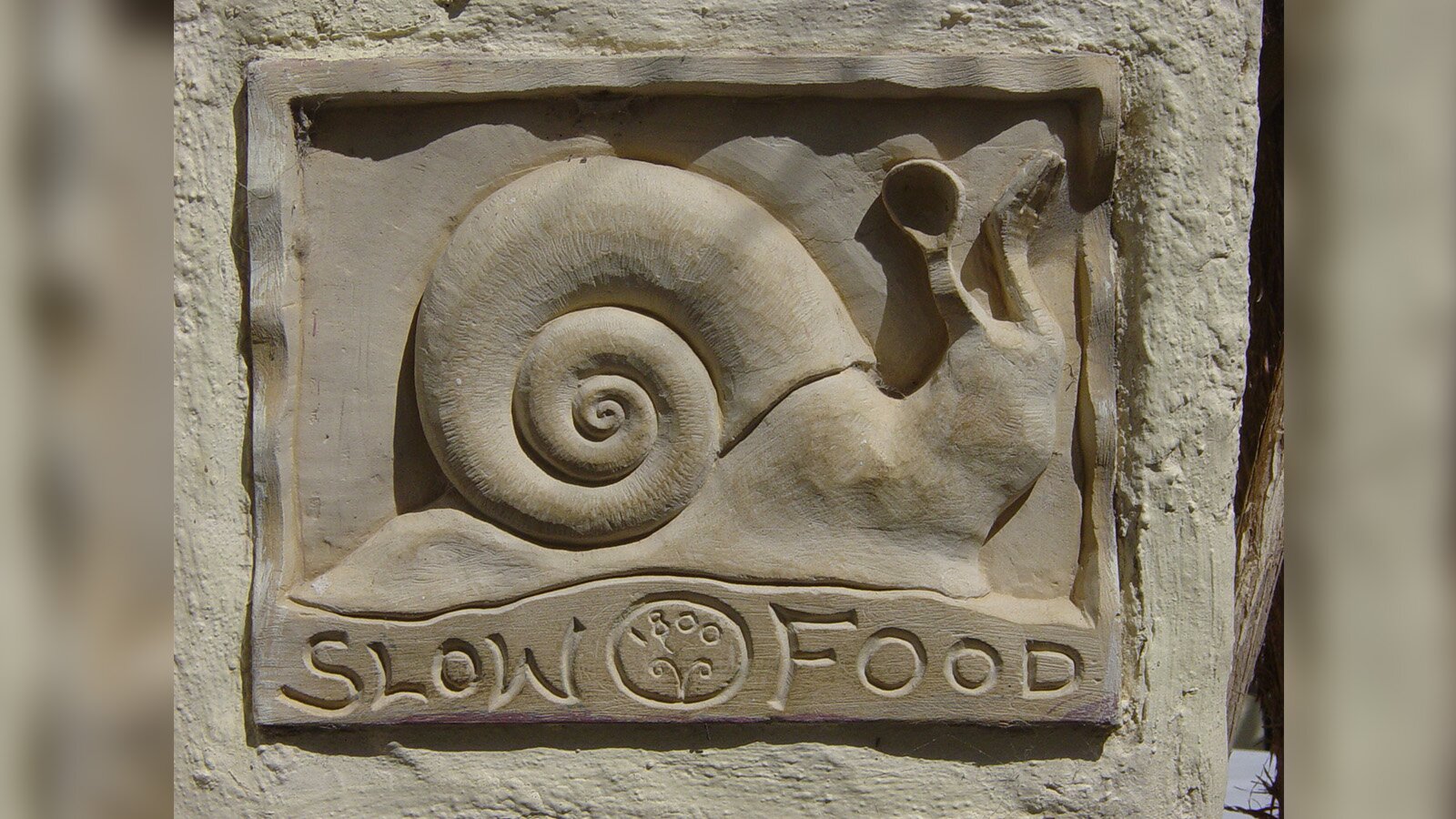 Zdjęcie przedstawia płaskorzeźbę z drewna, na której przedstawiony jest ślimak. Pod nim widoczny jest napis w języku angielskim: SLOW FOOD.