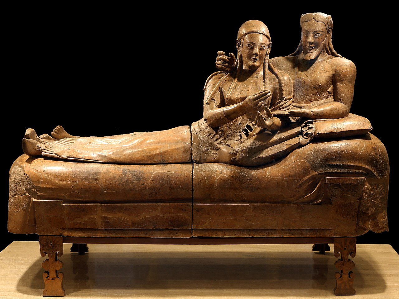 Ilustracja przedstawia sarkofag z rzeźbami kobiety i mężczyzny w pozycji półsiedzącej. Mężczyzna ma nagi tors, długie włosy z przedziałkiem na środku i brodę, opiera się na łokciu. Przed nim leży kobieta, ubrana w długą szatę, włosy zaplecione z kilka, małych warkoczyków.