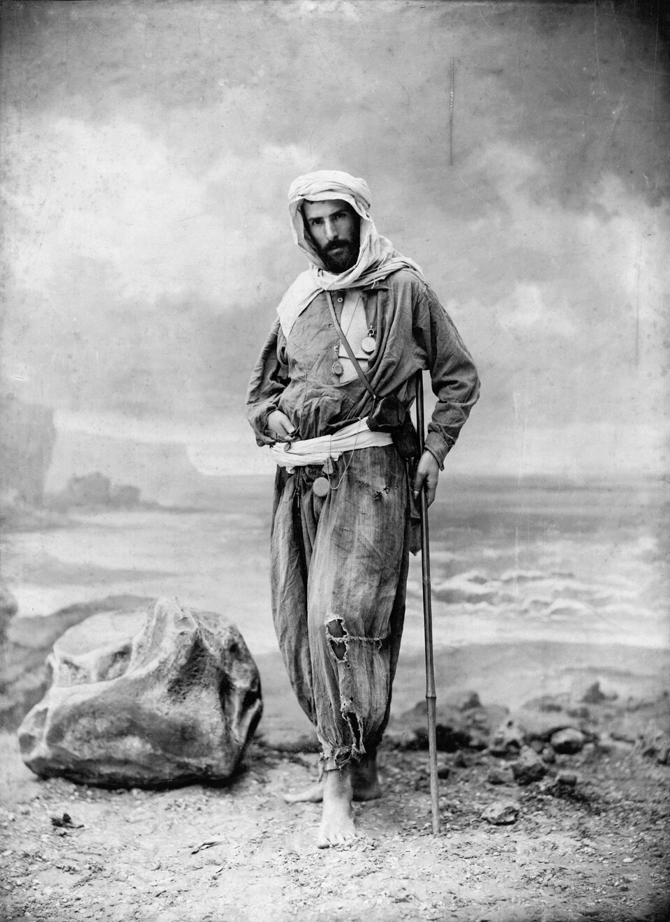 Czarno‑biała fotografia przedstawiająca brodatego mężczyznę stojącego boso na kamienistej ziemi. Ma na głowie turban lub owiniętą chustę i ubrany jest w luźny, długi strój podróżny przewiązany pasem. Na szyi i pasie widać kilka medalionów lub przyrządów, a przez ramię przewieszony jest pasek z małą torbą lub kaburą. Mężczyzna opiera się na lasce. W tle znajduje się pustynny krajobraz oraz duży kamień po lewej stronie.
