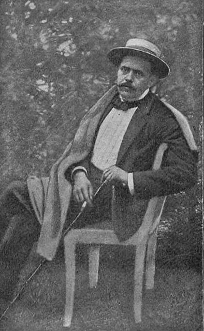 Zdjęcie przedstawia portret mężczyzny w średnim wieku. Mężczyzna ma pociągłą twarz. Cienie pod oczami. Ma starannie ostrzyżoną brodę i niewielkie wąsy. Ubrany jest w garnitur. Na głowie ma kapelusz.
