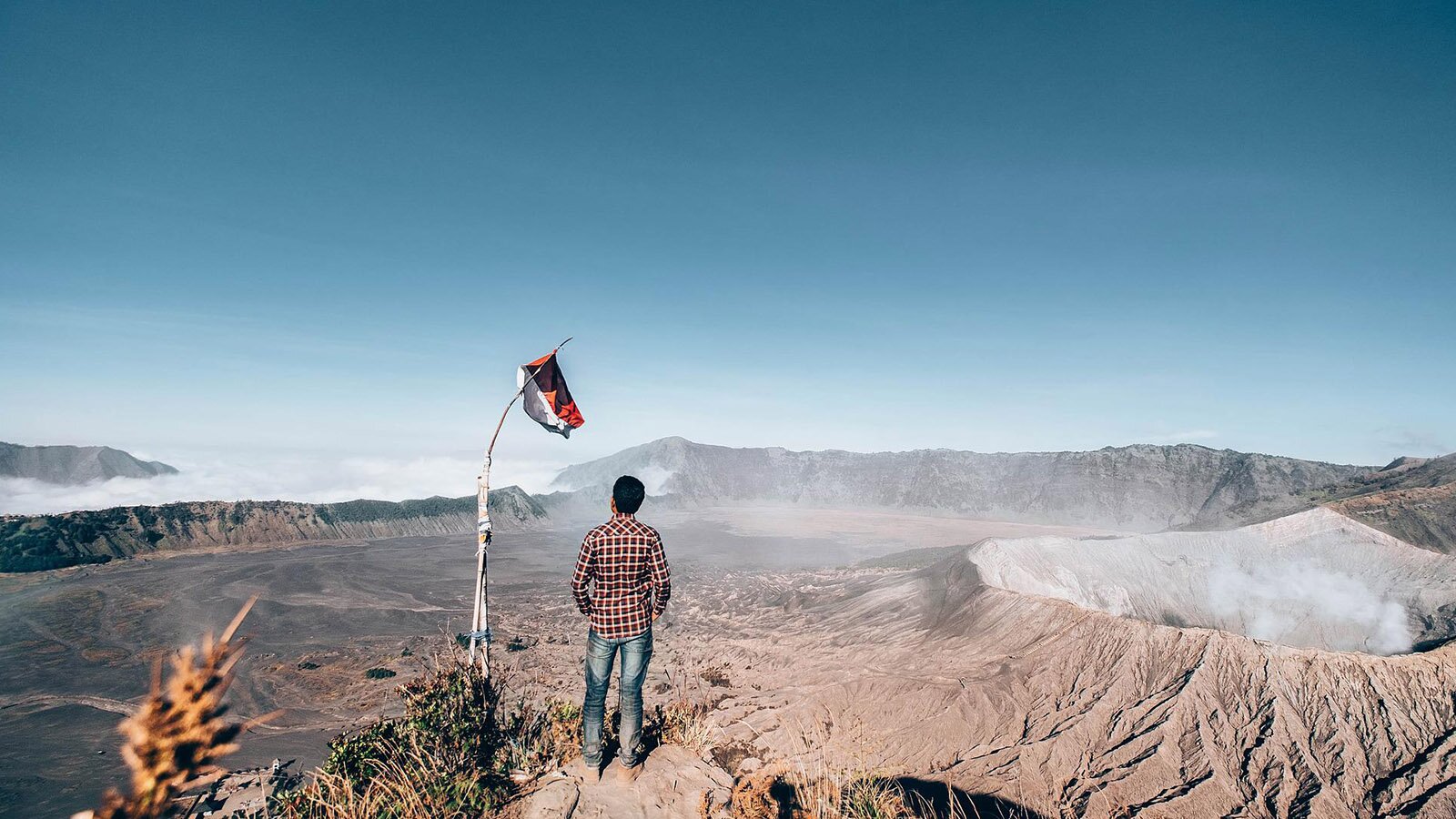 Zdjęcie przedstawia mężczyznę stojącego tyłem do obiektywu, przed nim rozpościera się pusta przestrzeń, nie zakłócona żadną roślinnością, jest tam tylko ciemna, sucha ziemia. W oddali ukazano niskie wzgórza. Po prawo od mężczyzny jest dymiący się krater wulkanu. 