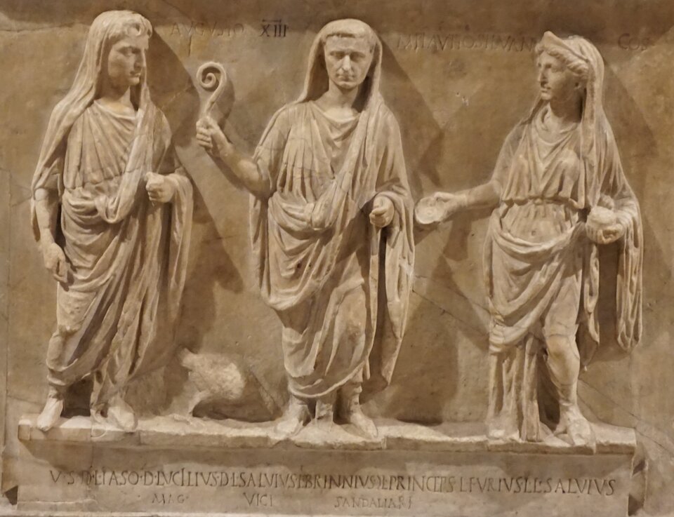 Zdjęcie przedstawia płaskorzeźbę wykonaną w kamieniu. Prezentuje ona trzy postacie. W środku stoi mężczyzna. Unosi rękę, w której trzyma przedmiot przypominający zawiniętą, jak róg, końcówkę pastorału.