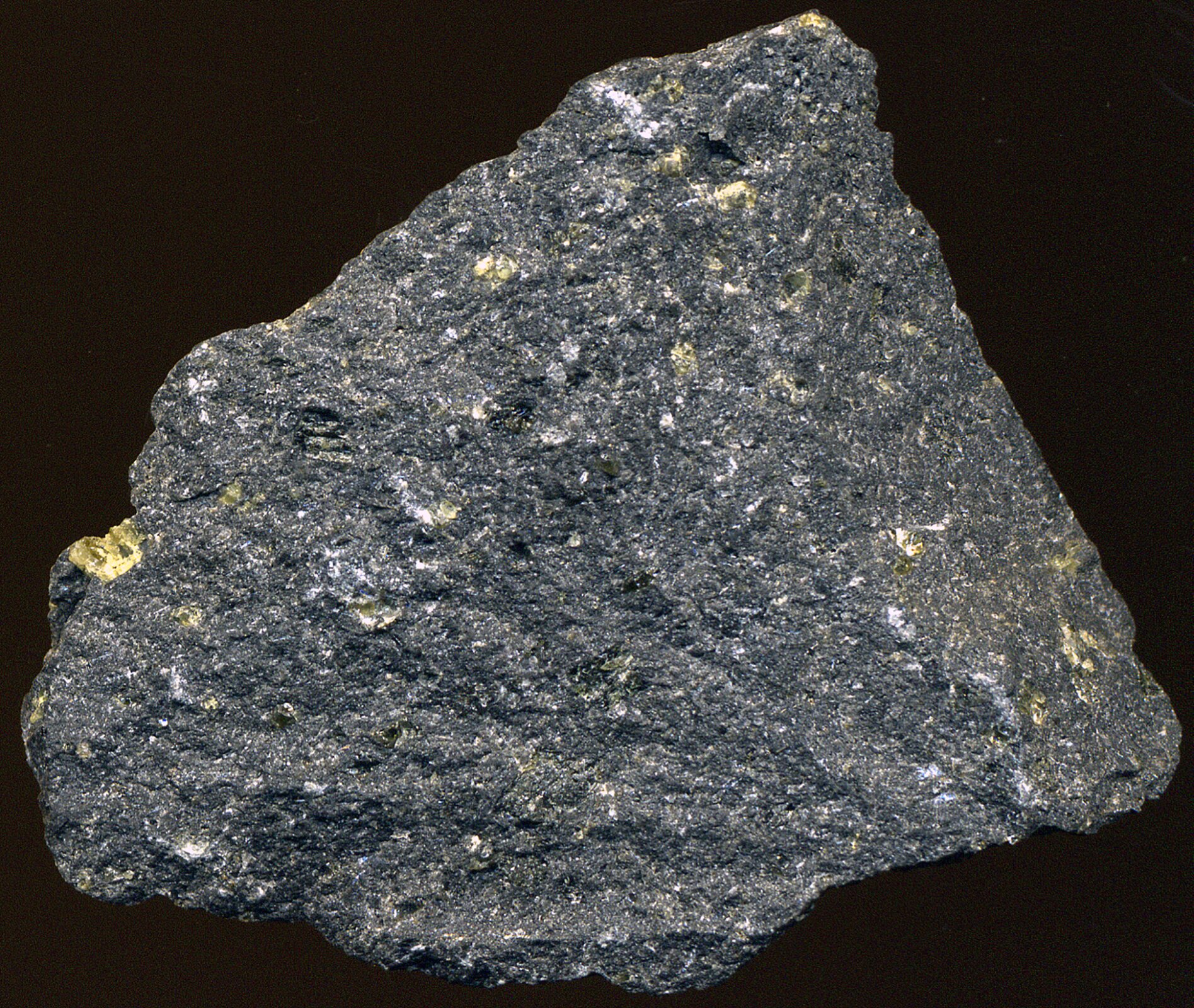 Fotografia prezentuje bazalt. Jest on ciemno‑szary z prześwitami białego i żółtego koloru. Jego kształt jest nieregularny, brzegi chropowate. 