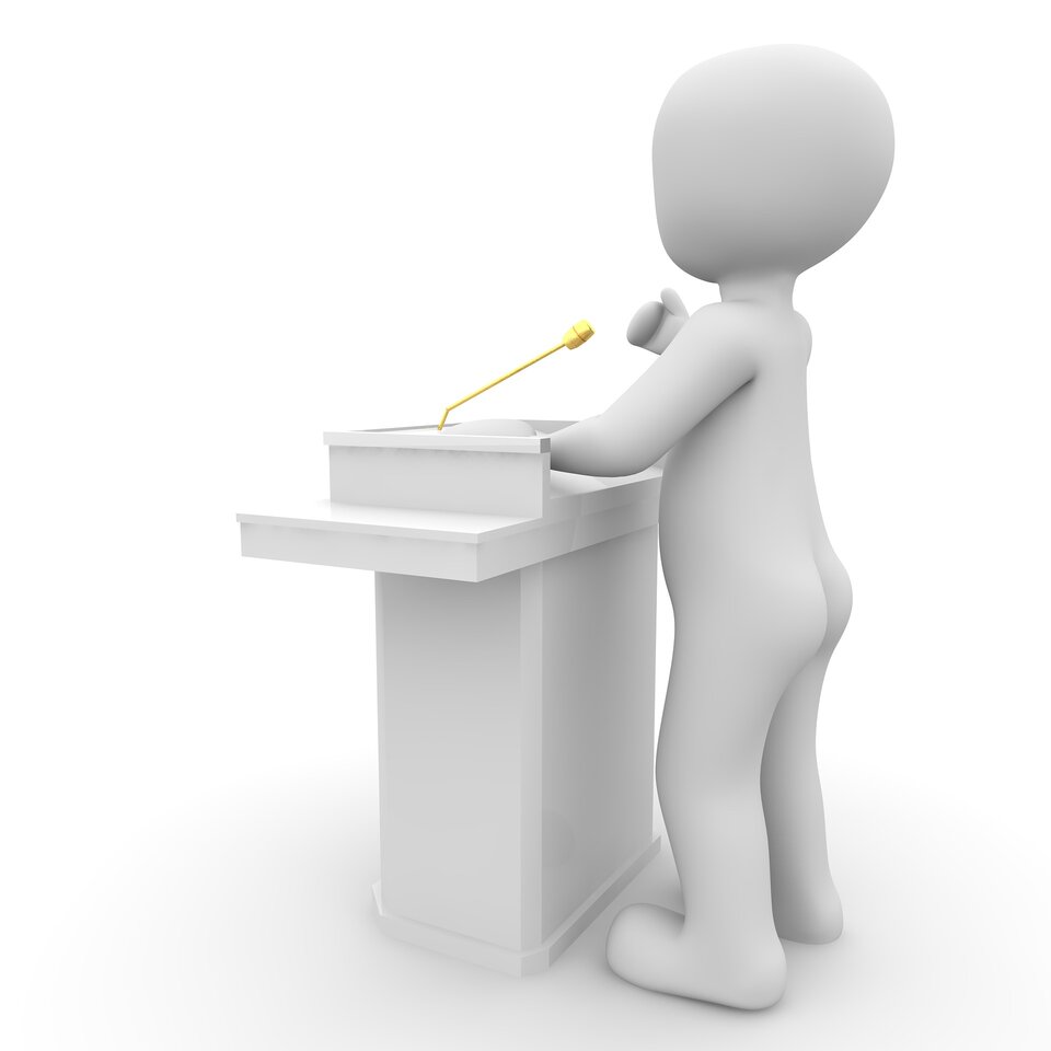 Ilustracja przedstawia białego ludzika stojącego przed białą mównicą. Scenę widzimy z profilu.