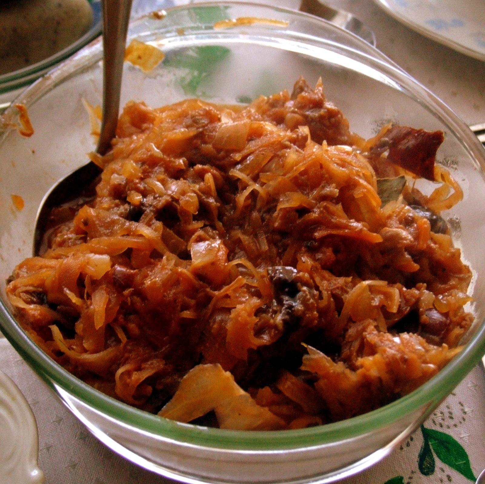 Zdjęcie przedstawia szklaną miskę z bigosem, czyli duszoną kapustą z mięsem i przyprawami. W miskę wetknięta jest łyżka.