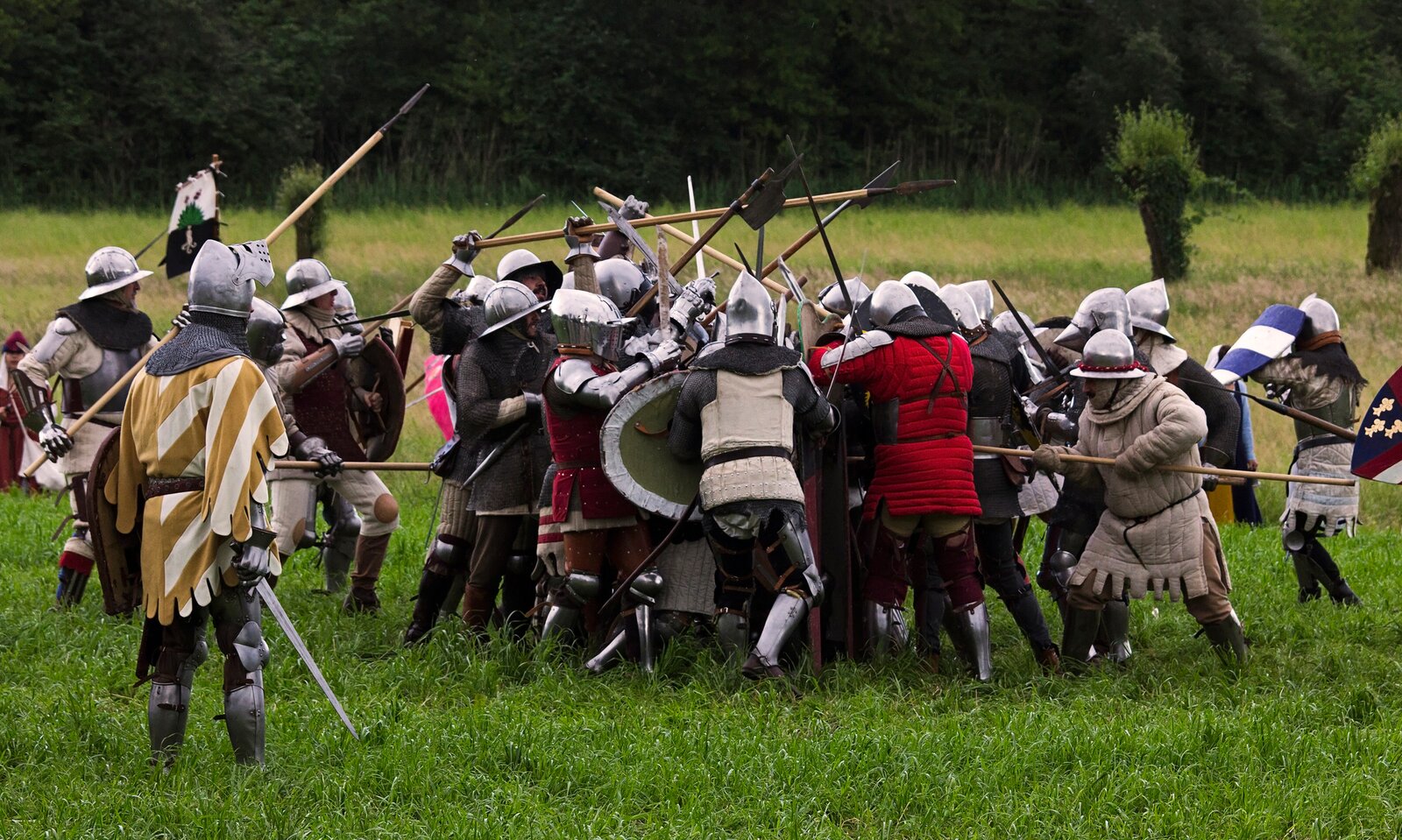 Zdjęcie przedstawia współczesną grupę rekonstrukcyjną. Mężczyźni walczą na polanie. Wielu z nich trzyma włócznie, inni miecze. Wielu ma także tarcze. Ubrani są w kaftany i zbroje oraz metalowe hełmy. Po lewej stronie stoi mężczyzna w zbroi, na którą ma nałożoną szatę w żółto‑białe pasy; w prawej ręce trzyma opuszczony miecz; przygląda się scenie walki.