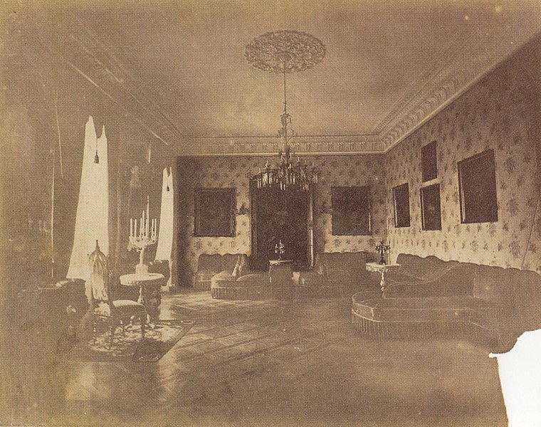 Zdjęcie przedstawia wnętrze dużego salonu. Stoi w nim duża kanapa, są także liczne fotele, stolik z dwoma krzesłami. Na wytapetowanych ścianach wiszą liczne obrazy. Pod sufitem wisi duży żyrandol.