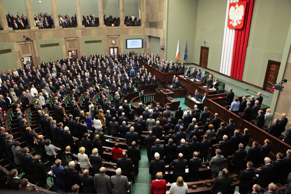 Zdjęcie przedstawia salę obrad polskiego Sejmu. Sala jest wypełniona posłami, którzy stoją. Sala ma kształt półkola - tu znajdują się ławy poselskie. Na końcu sali, na podwyższeniu zasiada Marszałek Sejmu, a po jego prawej stronie Rada Ministrów. Przed stołem Marszałka stoi mównica dla posłów.