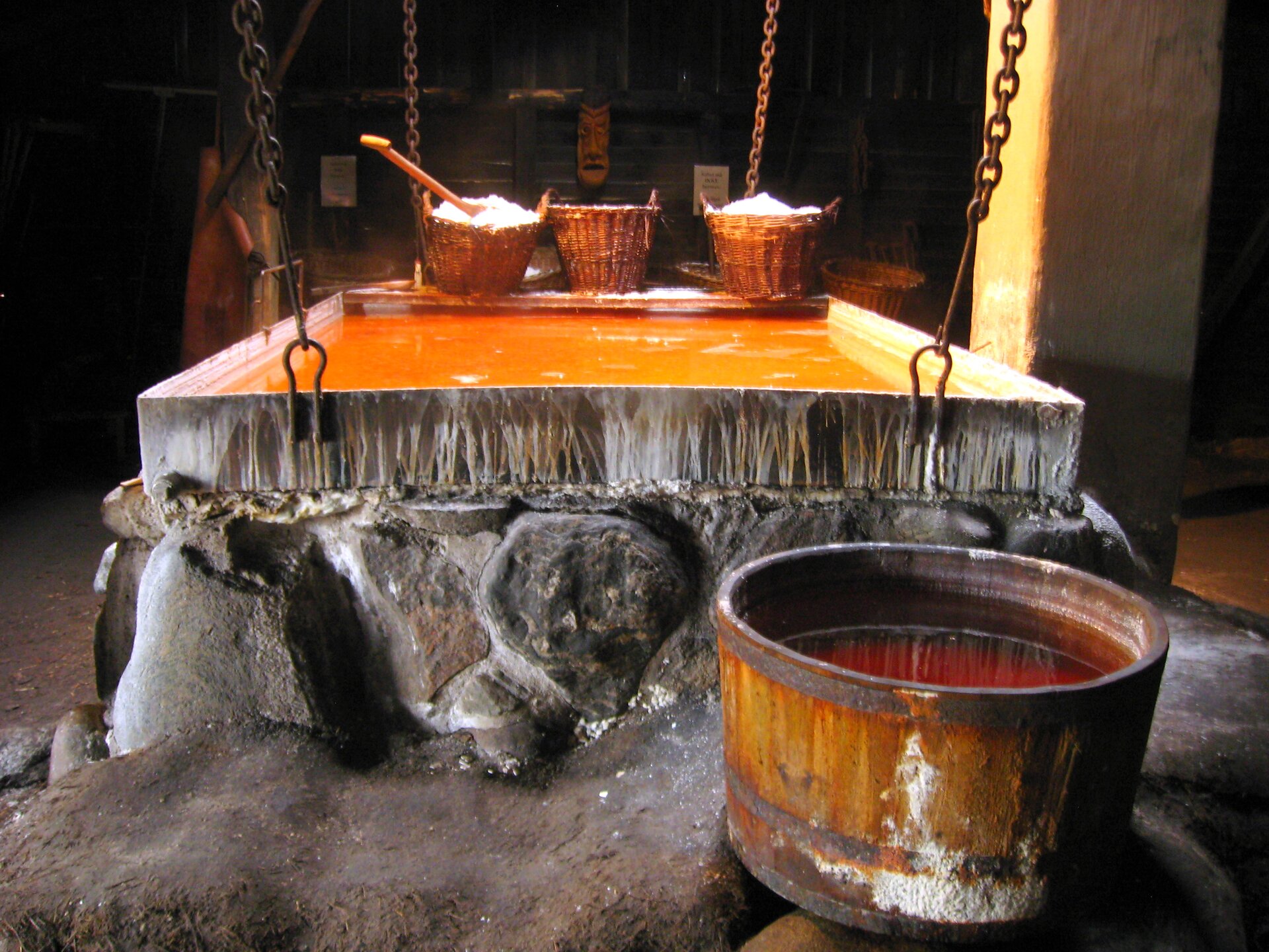 Zdjęcie przedstawia warzelnię soli. Ukazane jest prostokątne duże, płaskie naczynie stojąca na kamiennej podstawie. W naczyniu jest woda o pomarańczowej barwie. Widoczne są wiklinowe kosze wypełnione białą solą.