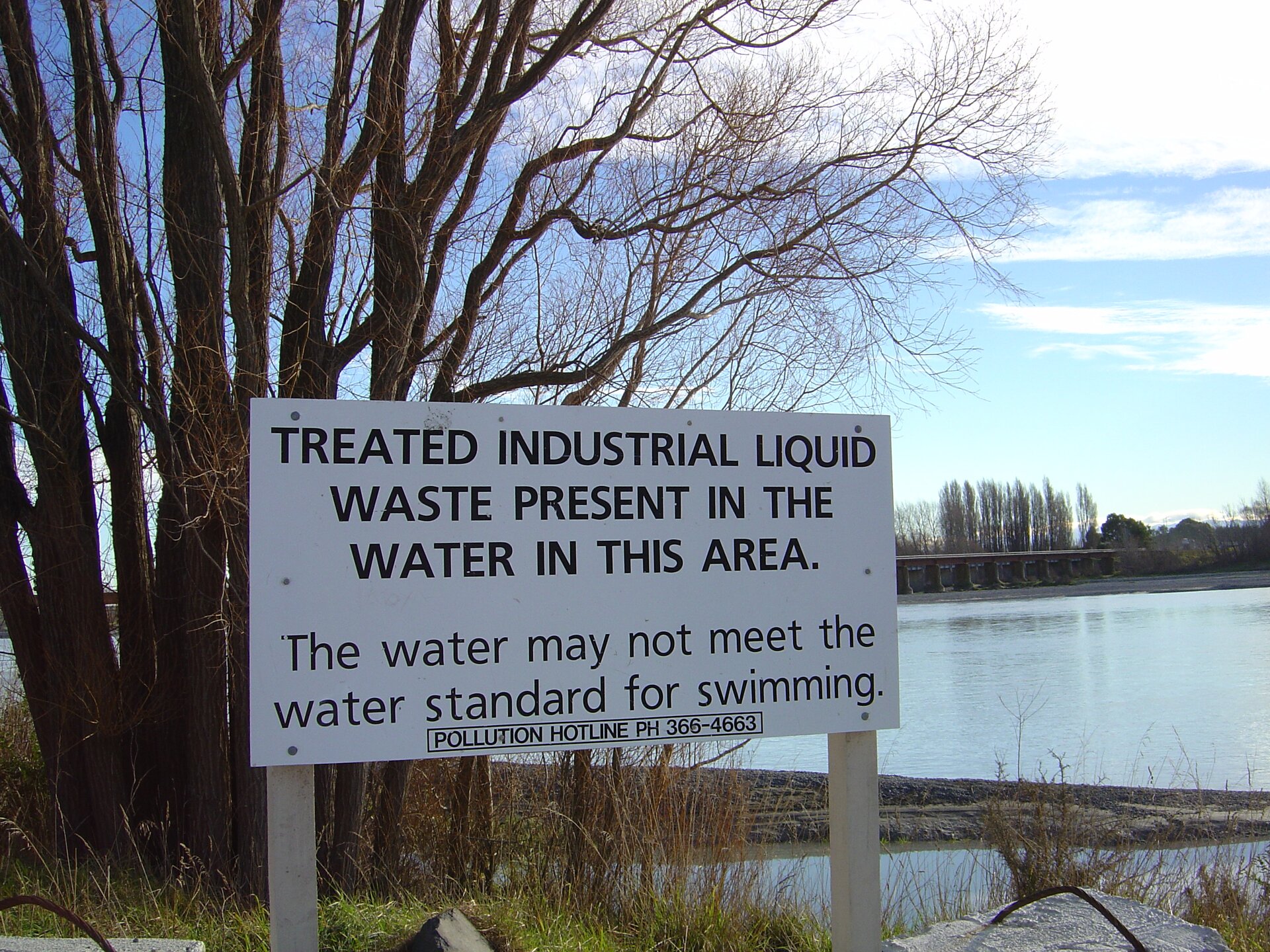 Na zdjęciu brzeg rzeki, krzewy. Na pierwszym planie tablica informacyjna. Czarny napis na białym tle. Informuje o zanieczyszczeniu wody.