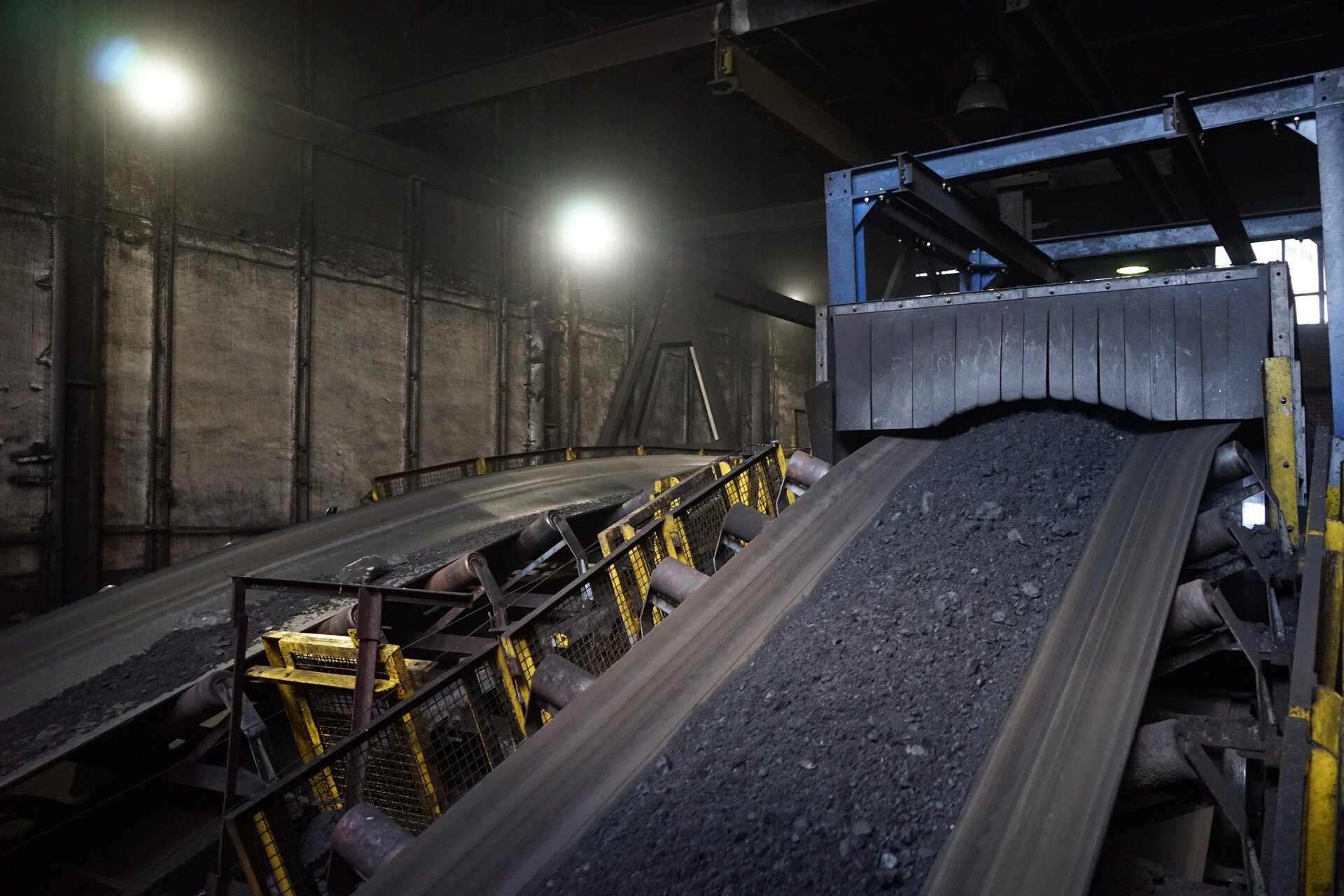 Na zdjęciu widoczne są dwa przenośniki Przenośniki widoczne na zdjęciu są układami modułowym. Na stalowej prostopadłościennej ramię osadzone są lekko nachylonych rolki. Rolki mają kształt walca. Na rolkach osadzona jest taśma. Na taśmie przenośnika widocznego po prawej stronie znajduje się pokruszony węgiel. Boki taśmy są zabezpieczone kratą oczkową uniemożliwiającej wypadnięcie urobku z taśmy. Na górze zdjęcia widać końcówkę taśmociągu. Taśma wchodzi pod gumowe zwisające paski. Oba przenośniki znajdują się pod kątem. Widoczne urządzenia znajdują się w budynku przemysłowym.Poniżej zdjęcia widoczny jest przycisk multimedialny z nagraniem audio. Treść nagrania jest zgodna z tytułem zdjęcia.