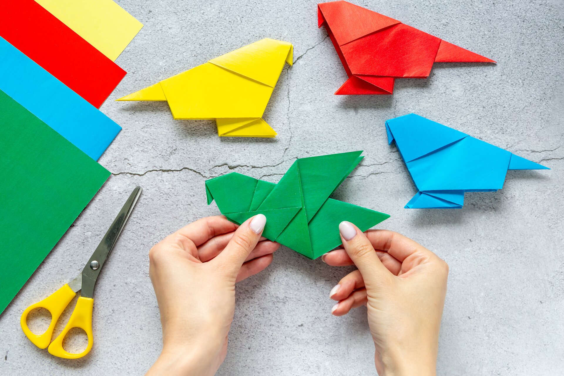 Zdjęcie przedstawia spękany blat stołu w kolorze szarym a na nim leżące cztery kartki papieru w kolorach: zielonym, czerwonym, niebieskim i żółtym. Obok kartek leżą nożyczki. Widoczne są też dłonie składające zielonego żurawia metodą origami. Na stole znajdują się też gotowe trzy żurawie w kolorach żółtym, niebieskim i czerwonym. 