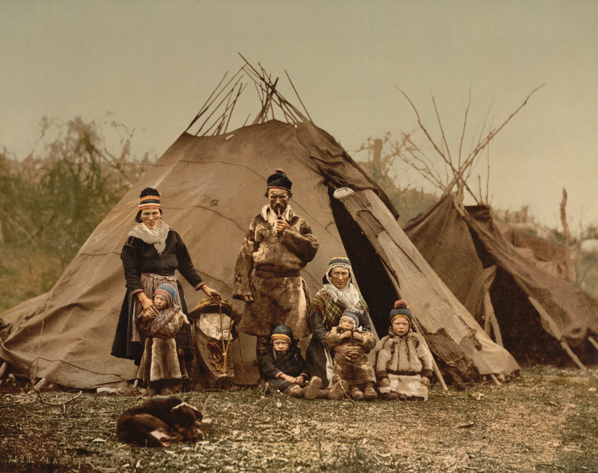 Fotografia przedstawia grupę osób w różnym wieku oraz psa. Są ubrani w ubrania ze skór i futer, a na głowach mają ludowe nakrycia. Kobieta z lewej strony przytrzymuje stojące przed nią dziecko. Po środku stoi mężczyzna palący fajkę. Kobieta po prawej klęczy na ziemi. Przed nią siedzi troje dzieci. Znajdują się na tle namiotu. Przed nimi leży zwinięty w kłębek pies. W tle widoczny jest jeszcze jeden namiot i krzaki.