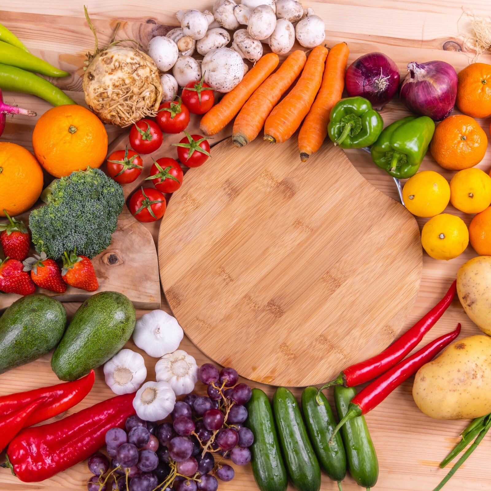 Zdjęcie przedstawia warzywa i owoce leżące na drewnianym blacie, ułożone wokół drewnianej deski. Widoczne są małe pomidory koktajlowe, marchew, czosnek, brokuł, czerwona i zielona papryka, ogórek, czerwona cebula, ziemniaki, seler korzeniowy, pieczarki, awokado oraz pomarańcze, truskawki, winogrona, cytryny i mandarynki. 