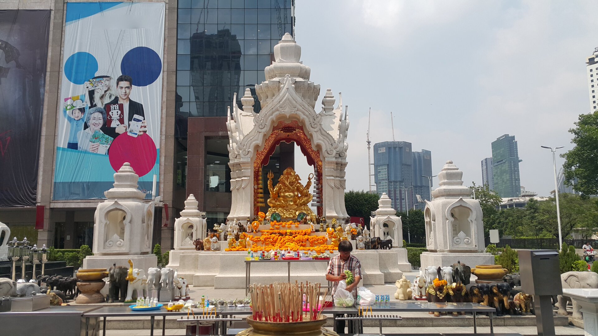 Zdjęcie przedstawia plac znajdujący się przed centrum handlowym, na którym widoczna jest świątynia poświęcona świętej trójcy hinduizmu. Pośrodku znajduje się duża zdobiona konstrukcja, ze złotym posągiem bóstwa, umieszczony na podwyższonym cokole, pod małym baldachimem. Na czterech narożnikach widoczne są mniejsze kapliczki zbudowane w tym samym stylu. Przed posągiem widoczne są kadzidła, girlandy kwiatowe i świece. W tle widoczna jest przeszklona fasada wielopiętrowego budynku z ogromnym bilbordem przedstawiającym dwie kobiety – starszą i młodą, młodego mężczyznę i dziecko, którzy reklamują różne produkty.