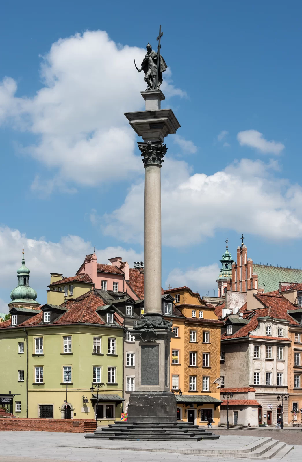 Fotografia przedstawia kolumnę umieszczoną na wysokim cokole, na której szczycie stoi figura mężczyzny w średnim wieku ubranego w pełną zbroję, w jednej dłoni trzyma szablę, a w drugiej wysoki krzyż. Na głowie ma koronę, na której umieszczone jest jabłko z krzyżem. Kolumna stoi na placu, za nią widać kolorowe kamienice.
