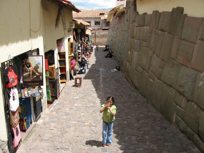 Kliknij, aby powiększyć. Na zdjęciu ulica. Z lewej budynki ze sklepami: część kolorowego towaru (torby, ubrania, obrazy) została wystawiona na ulicę. Z prawej ściana budynku, która w dolnej części obłożona jest płaskimi, prostokątnymi kamieniami. Na środku ulicy stoi dziewczynka w czarnych włosach. Ubrana jest w niebieskie spodnie i zieloną koszulkę z krótkim rękawkiem. Przykłada obie dłonie do ust. W tle, pod murem, siedzą dwaj mężczyźni. Na końcu ulicy mur, a za nim budynek mieszkalny.