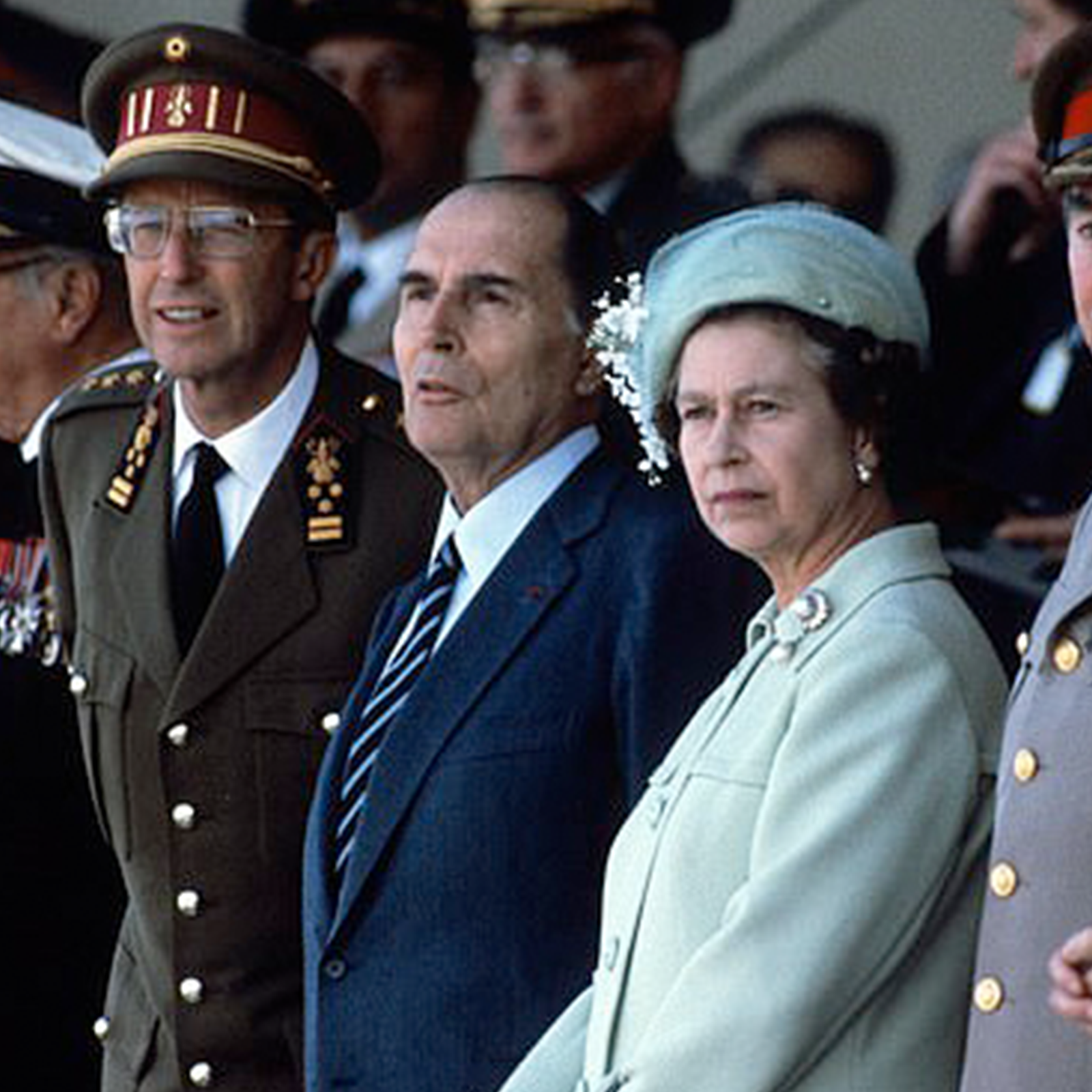 Zdjęcie przedstawia królową Elżbietę II uczestniczącą w jakimś wydarzeniu. Widoczna jest kobieta w średnim wieku, ubrana w jasnoniebieski płaszcz i jasnoniebieski kapelusz. Zwrócona jest w lewo i patrzy przed siebie. Obok niej widoczni są mężczyźni w starszym wieku, w tym jeden ubrany ma ciemnogranatowy garnitur, a drugi w mundur. 