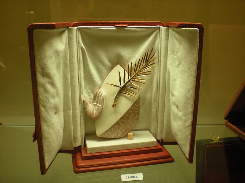 Zdjęcie przedstawia nagrodę w postaci owalnej figurki z otwartą dłonią, napisem i złotym liściem palmowym. Znajduje się ona w otwartym futerale wyłożonym jasnym welurem. Przed nim stoi mała kartka z napisem Cannes.