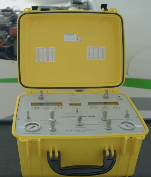 Zdjęcie przedstawia Tester PS525. Widoczna jest otwarta żółta skrzynka z czarnym uchwytem z boku. Wewnątrz skrzynki widoczne są w dwóch rogach wskaźniki zegarowe. Pomiędzy wskaźnikami znajdują się przełączniki. Ponad przełącznikami widoczne są trzy małe wyświetlacze cyfrowe.