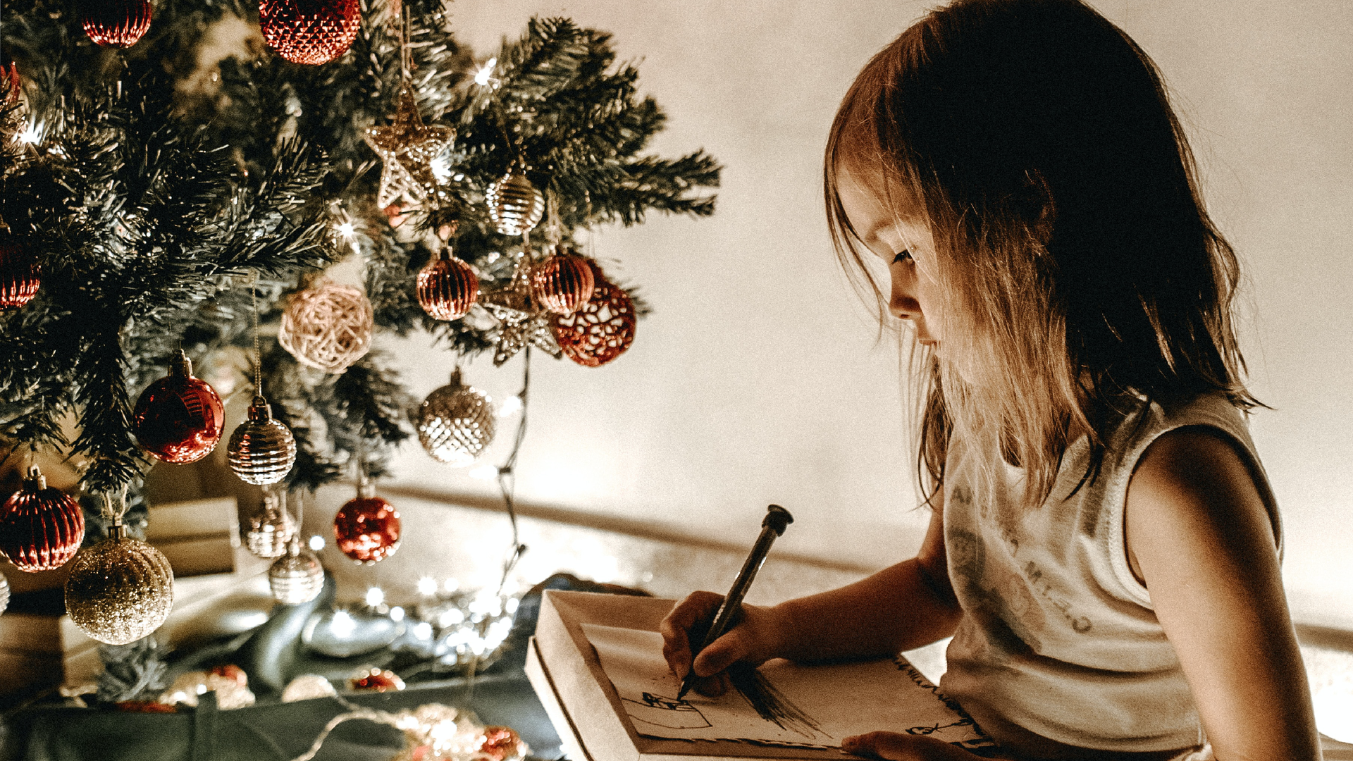 Zdjęcie przedstawia małą dziewczynkę, która coś rysuje na papierze. Obok dziewczynki znajduje się choinka.