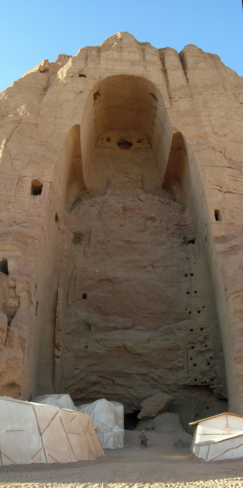 Na zdjęciu znajduje się wnęka skalna, w której znajdował się posąg Buddy. Znajdują się w niej jeszcze pewne jego śladu. We wnęce znajdują się liczne otwory. Pod skałą znajdują się namioty oraz mężczyzna w pomarańczowym kasku zmierzający w stronę wnęki. 