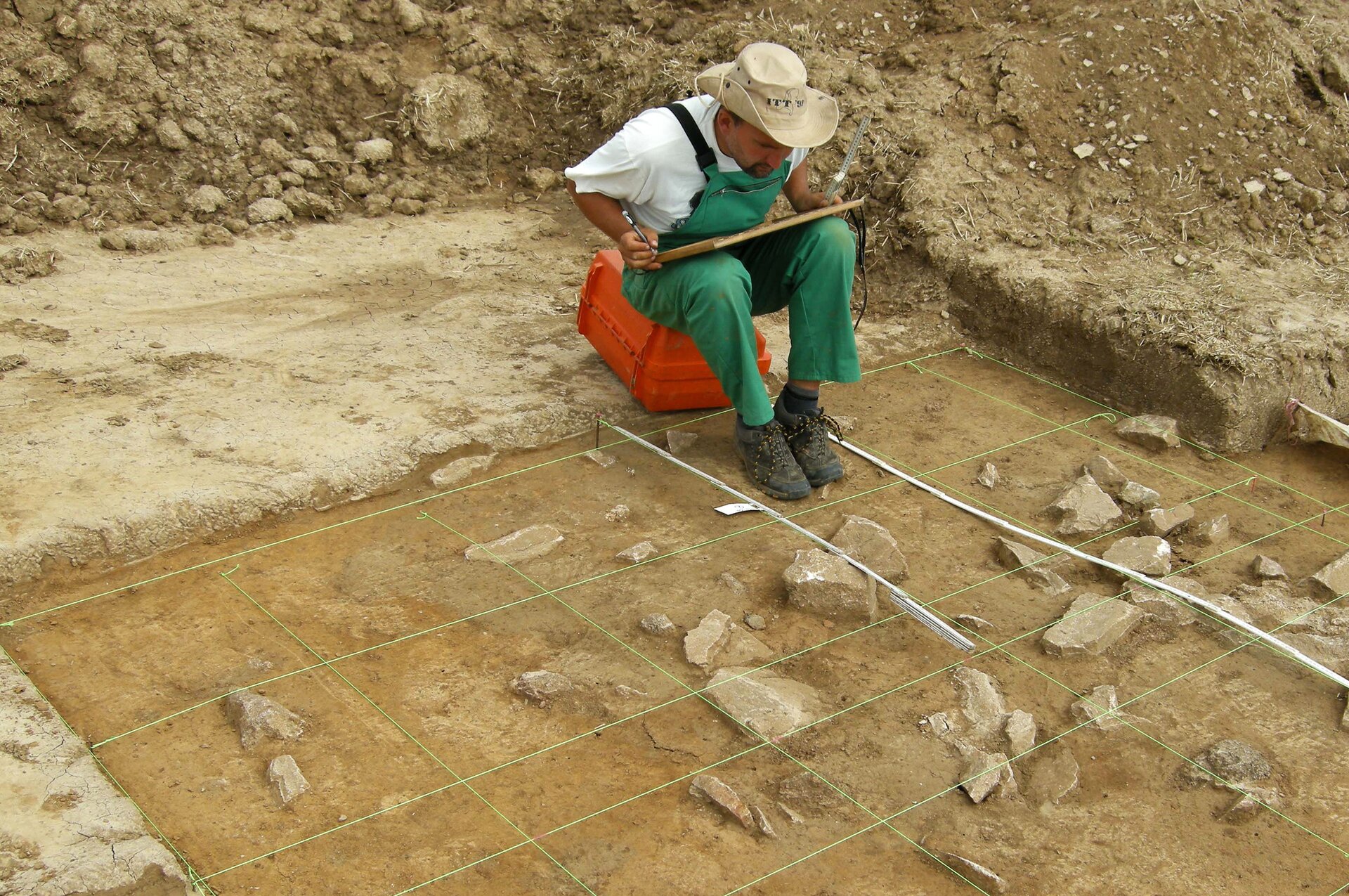 Zdjęcie przedstawia archeologa siedzącego w dole ze znaleziskiem, rysującego i opisującego leżące przed nim ślady archeologiczne.