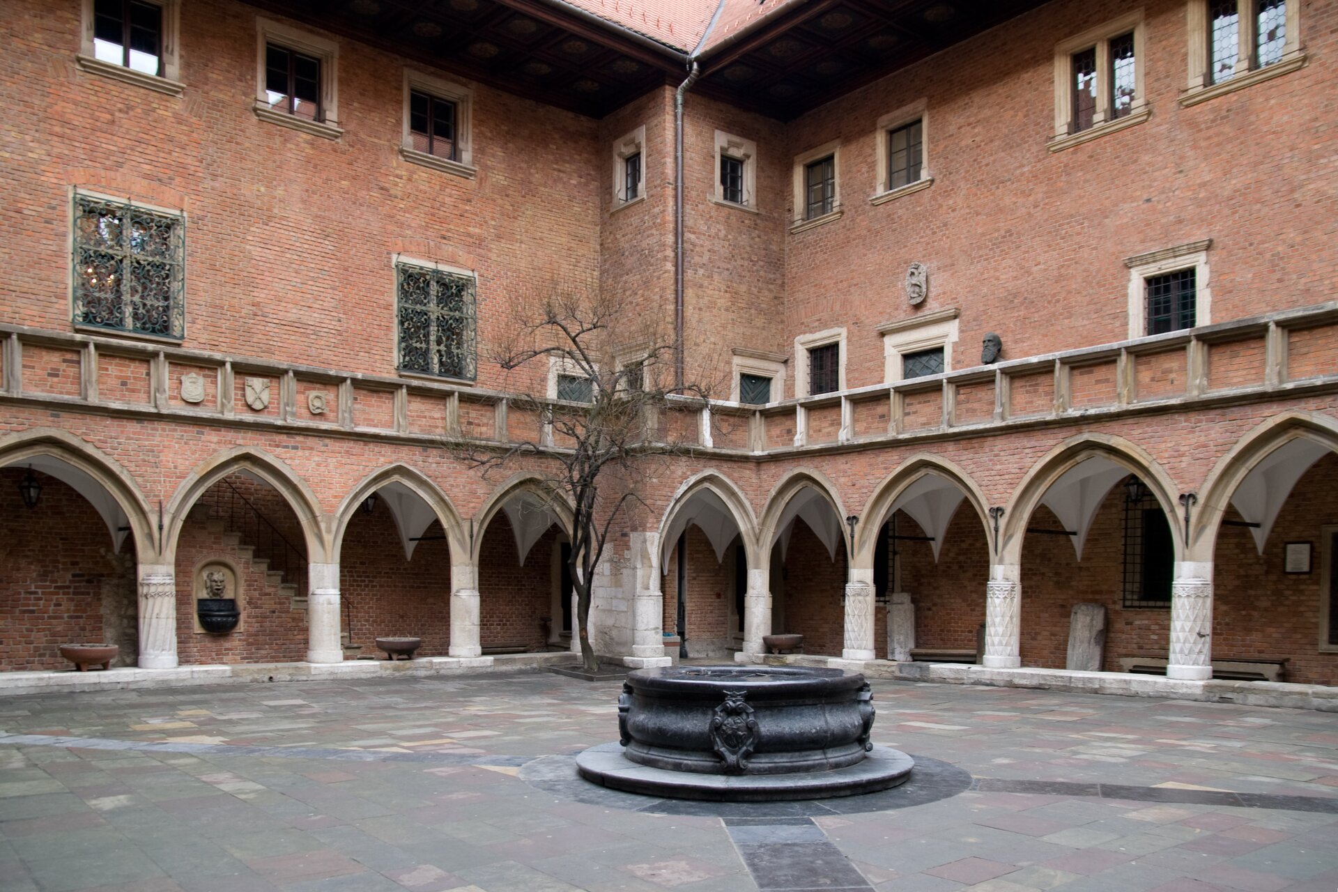 Ilustracja przedstawia Collegium Maius. To budowla zbudowana z cegły. Jest wzniesiona na arkadach. Znajdują się krużganki. Powyżej umiejscowiona jest loggia. Okna mają kształt prostokątny. Na środku dziedzińca znajduje się studnia.