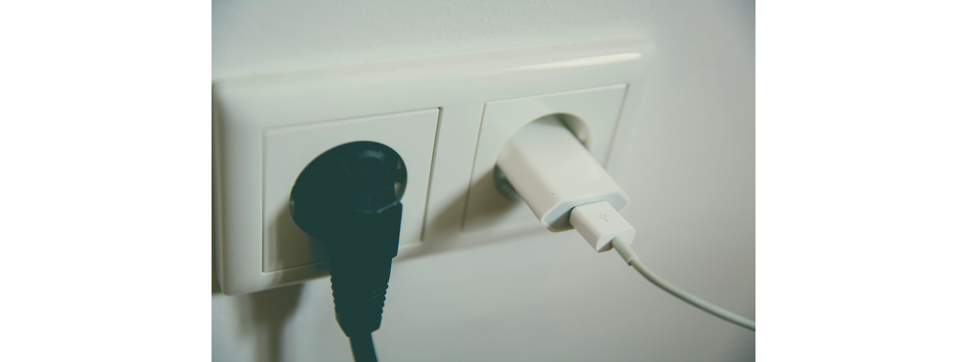 Zdjęcie przedstawia dwa gniazdka elektryczne w kremowym kolorze zamontowane w jasnej ścianie. Do gniazdek podłączono wtyczki. Jedna wtyczka jest koloru białego, druga zaś- czarnego.