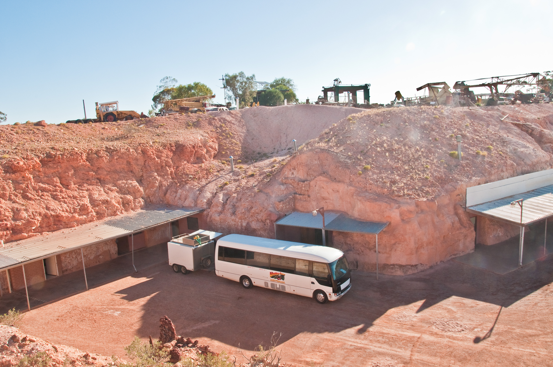 Fotografia prezentuje wyrobisko kopalni cennych kamieni. W ścianach wyrobiska znajdują się zadaszone wejścia do opuszczonych chodników kopalni. Przed jednym z wejść stoi biały autokar.