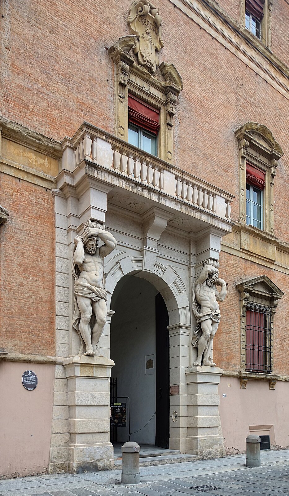 Zdjęcie przedstawia portal o łukowym zakończeniu, zbudowany z jasnego kamienia. Wbudowany został w murowany, nieco ciemniejszy budynek. Po obu stronach łukowego przejścia znajdują się rzeźby stojących mężczyzn. Mężczyźni są nadzy, przewiązani w pasie wyrzeźbioną chustą. Mają mocno zarysowane mięśnie, zmarszczone czoło i bujny zarost. Obie ręce trzymają przy twarzy. Nad nimi umieszczono balkon, który podtrzymują na swoich głowach.