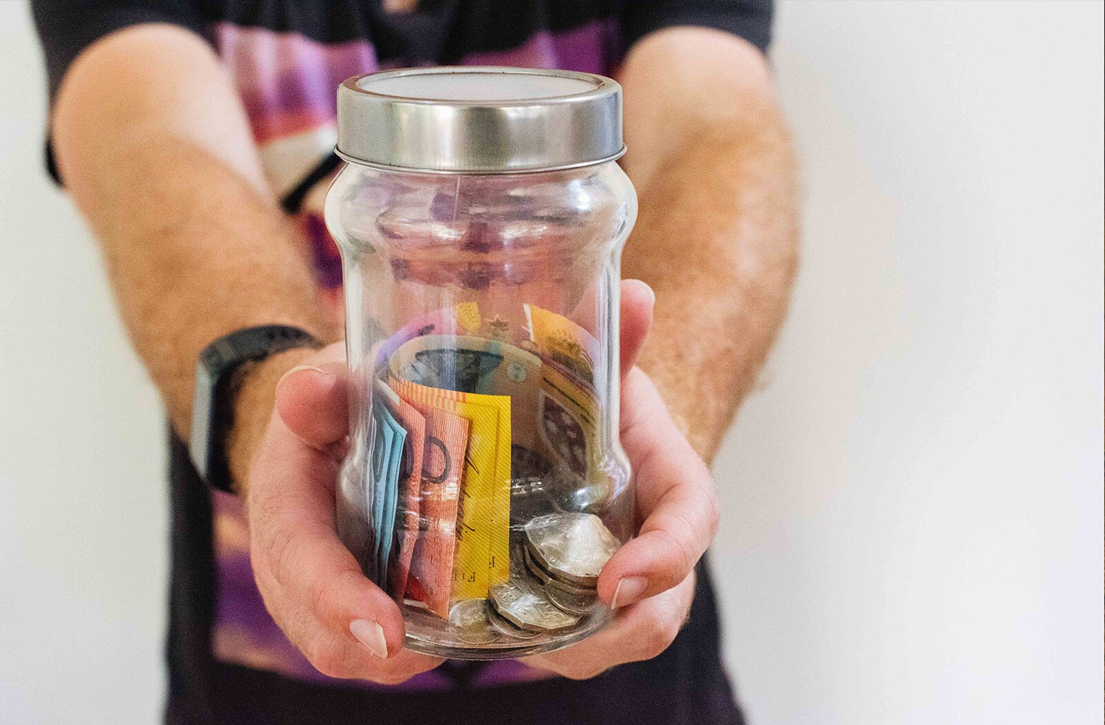 Zdjęcie przedstawia wyciągnięte do przodu ręce mężczyzny. W tle widoczny jest korpus postaci w kolorowej koszulce. Mężczyzna trzyma w dłoniach szklany słoik ze srebrną nakrętką. W słoiku widoczne są: plik różnokolorowych banknotów oraz monety.