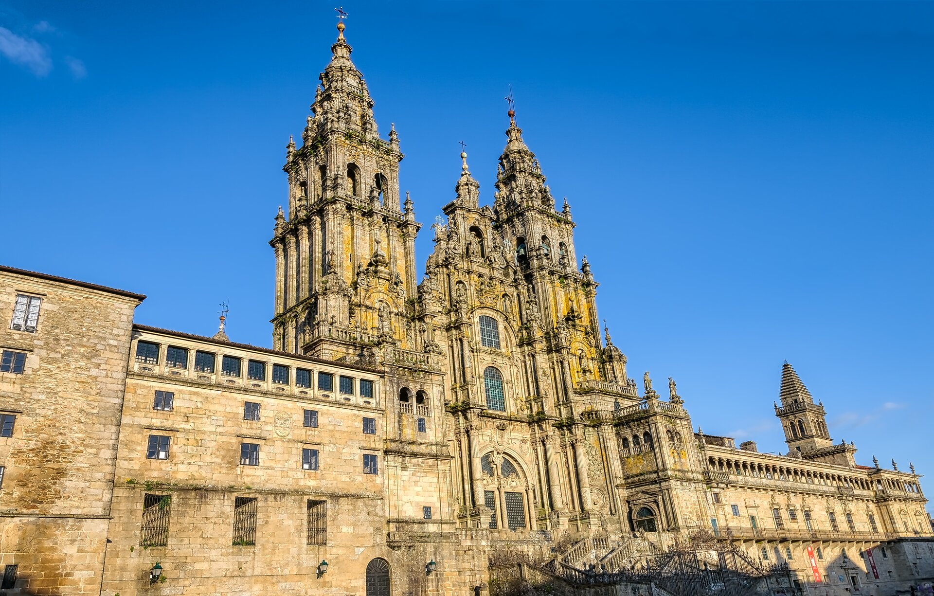 Ilustracja przedstawiająca katedrę w Santiago de Compostela w Hiszpanii. Dwupoziomowy kościół jest trójnawową bazyliką z szerokim transeptem oraz prezbiterium z półkolistą apsydą. Zachodnią fasadę znana jako Fachada de la Plaza del Obradoiro flankują dwie średniowieczne wieże wzbogacone obfitą dekoracją rzeźbiarską z XVIII wieku. Wzniesione na planie kwadratu, mają wysokość 76 metrów. Wejście główne do górnego kościoła poprzedza niewielki kwadratowy plac, do którego prowadzą ciągi schodów.