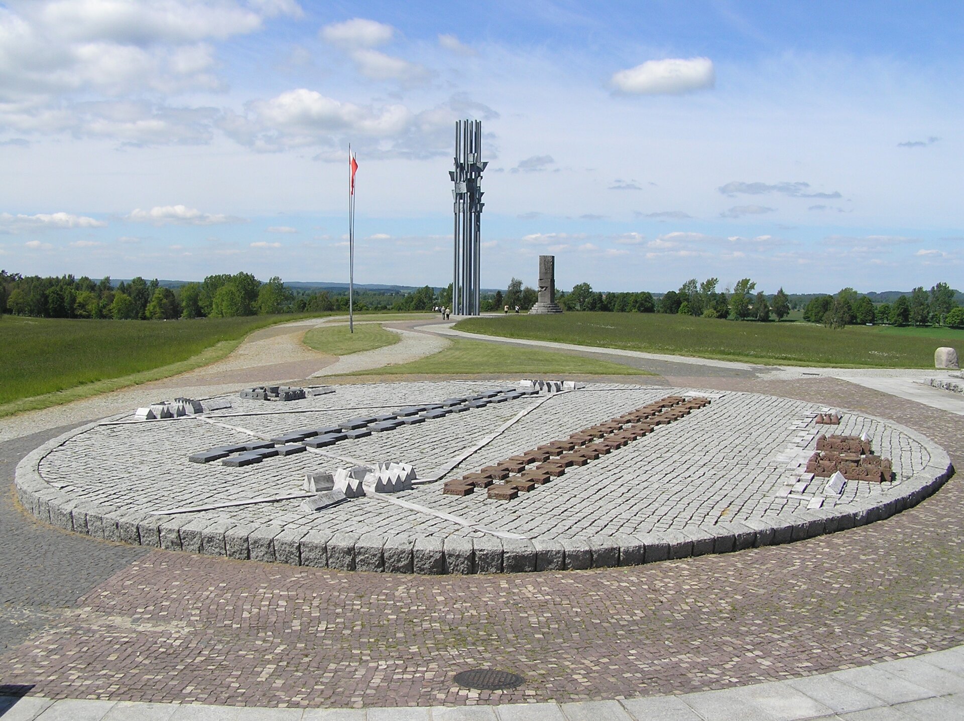 Na zdjęciu przedstawiona jest kamienna okrągła makieta ukazująca rozmieszczenie wojsk polskich i krzyżackich podczas bitwy pod Grunwaldem. W tle widoczny jest wysoki pomnik grunwaldzki i błękitne niebo. 