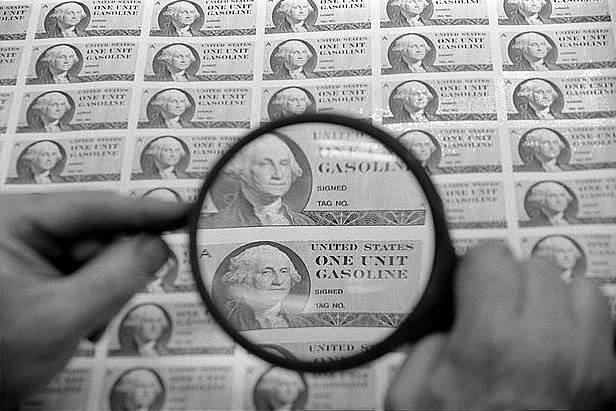 Fotografia przedstawia dwie dłonie trzymające lupę powiększającą. Pod lupą znajduje się zdjęcie mężczyzny z jasnymi włosami i kołnierzykiem białej koszuli. Obok widnieją napisy „UNITED STATES” oraz „ONE UNIT GASOLINE”. Wszystko razem jest na szarym tle w formie prostokąta. Takich prostokątów poza zasięgiem lupy jest wielokrotnie więcej. Wydrukowane są one na białym papierze. Cała fotografia jest czarno‑biała.