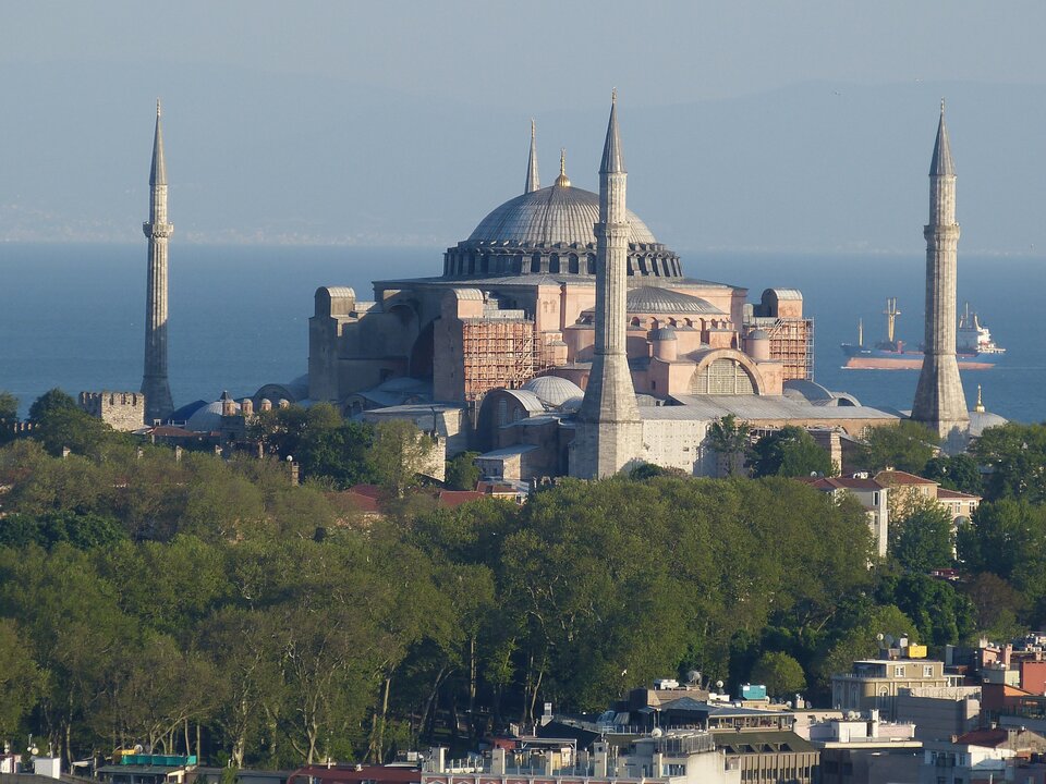 Zdjęcie współczesne, przedstawiające kościół Hagia Sophia w Stambule. Widoczna w centralnej części zdjęcia konstrukcja nawy głównej z charakterystyczną owalną kopułą, dookoła której symetrycznie w kształcie kwadratu ustawiono cztery minarety. W tle widoczne wody Morza Marmara, na którym po prawej stronie zdjęcia widać pływające statki. W dolnej części zdjęcia drzewostan i zabudowania domostw w Stambule. 