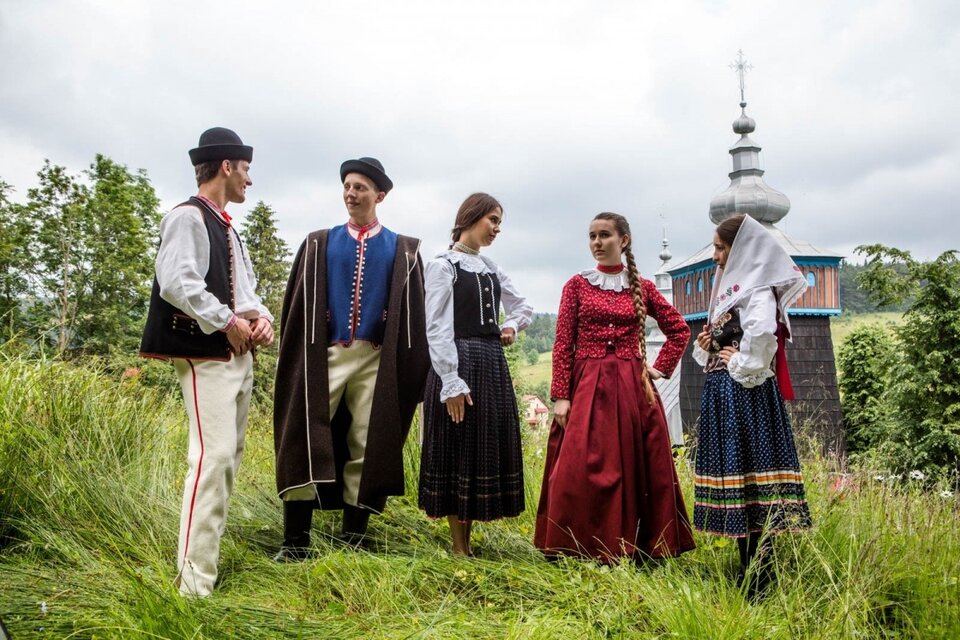 Zdjęcie przedstawia stojącą na łące młodzież ubraną w tradycyjne stroje łemkowskie. Panowie mają kremowe spodnie z czerwonymi lampasami, białe koszule oraz czarne lub niebieskie kamizelki z czerwoną obwódką, a na głowach okrągłe kapelusiki z niewielkim rondem. Kobiety ubrane są w bluzki z haftowanym, marszczonym kołnierzem. Dwie z nich mają białe bluzki, na to założone czarne kamizelki, trzecia czerwoną bluzkę. Mają długie, falbaniaste spódnice: dwie z nich są granatowe a jedna czerwona. Włosy kobiet splecione są w długi warkocz, jedna z dziewczyn ma na głowie białą chustę.